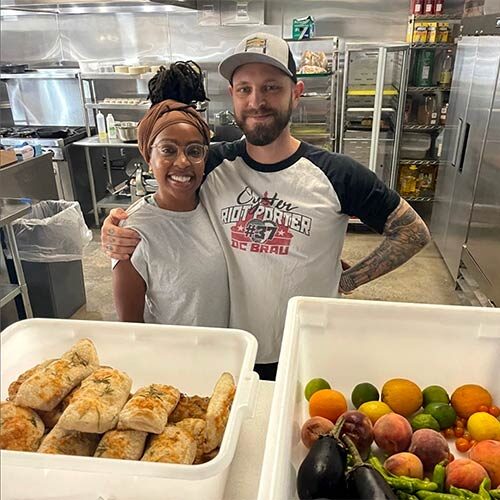 A man and woman stand together in an industrial kitchen with bins of fresh food in the foreground